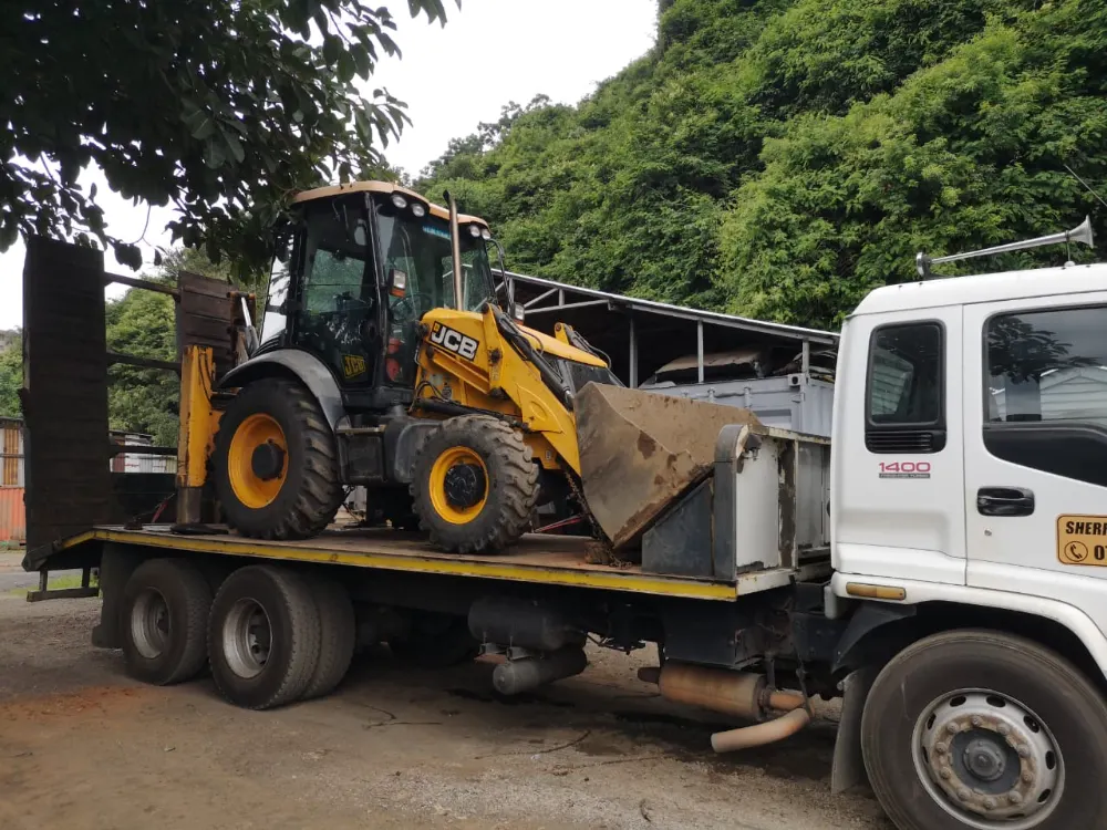 A JCB TLB loaded on Sherry's flatbed truck under the trees at the yard